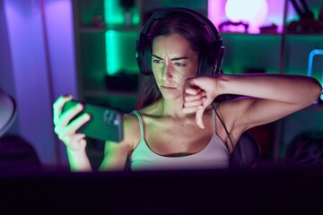 Young brunette woman playing video games with smartphone with angry face, negative sign showing dislike with thumbs down, rejection concept