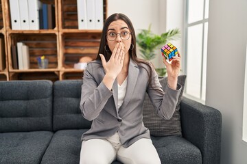 Hispanic woman playing colorful puzzle cube intelligence game covering mouth with hand, shocked and...