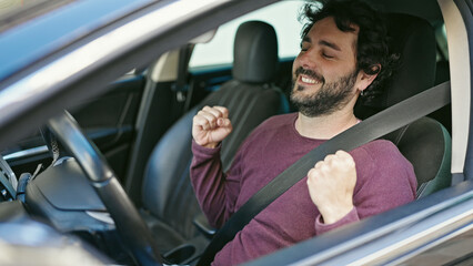 Young hispanic man sitting on car with winner expression at street