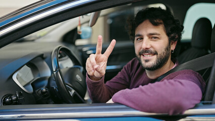 Young hispanic man sitting on car doing victory gesture at street - Powered by Adobe