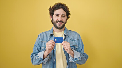 Young hispanic man holding credit card standing over isolated yellow background