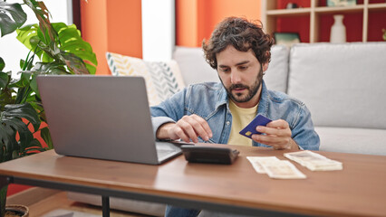 Young hispanic man using laptop and calculator counting denmark krone banknotes at home