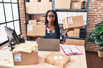 African woman with braids working at small business ecommerce holding packages puffing cheeks with funny face. mouth inflated with air, catching air. - obrazy, fototapety, plakaty