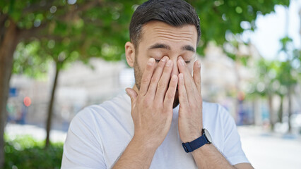 Young hispanic man standing with serious expression rubbing eyes at street