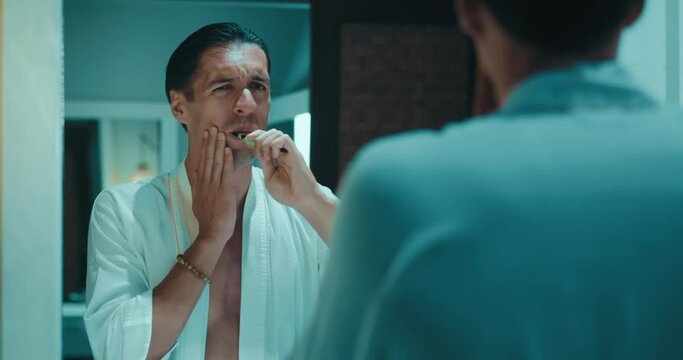 Reflection in the mirror young man in bathrobe brushing teeth with toothbrush in modern bathroom and stops because toothache. Concept of dental problems and toothache. Man suffering from teeth pain