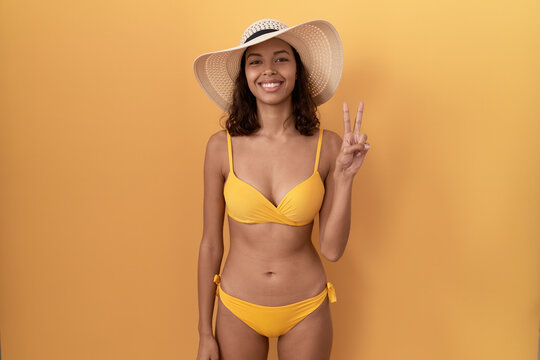 Young hispanic woman wearing bikini and summer hat showing and pointing up with fingers number two while smiling confident and happy.