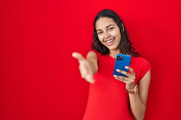 Young brazilian woman using smartphone over red background smiling friendly offering handshake as greeting and welcoming. successful business.