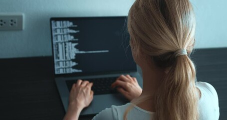 Back view young woman computer programmer looking at computer monitor and writing program code sitting at workplace in office. IT software developer typing data coding programming with laptop computer