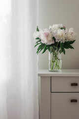 Bouquet of white peonies in a glass vase on a white wooden chest of drawers