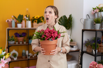 Young caucasian woman working at florist shop holding plant pot angry and mad screaming frustrated and furious, shouting with anger. rage and aggressive concept.