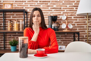 Young hispanic woman drinking coffee at home covering mouth with hand, shocked and afraid for...