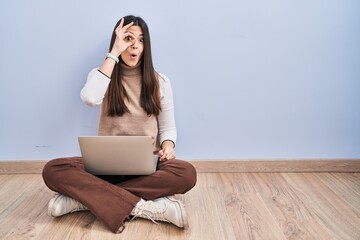 Young brunette woman working using computer laptop sitting on the floor doing ok gesture shocked with surprised face, eye looking through fingers. unbelieving expression.