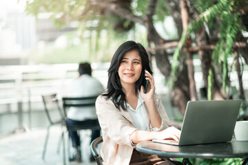 Attractive asian business woman using smartphone for talking with colleague while using laptop for work at co-working space, Urban business people, work anywhere lifestyles.