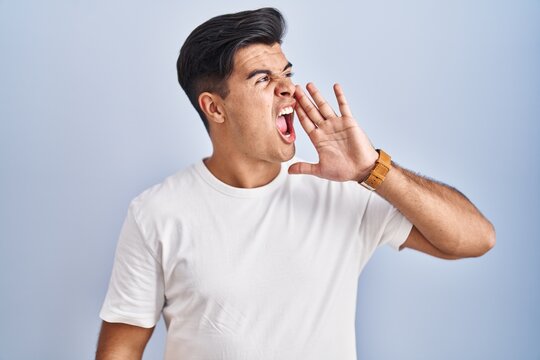 Hispanic man standing over blue background shouting and screaming loud to side with hand on mouth. communication concept.
