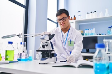 Down syndrome man wearing scientist uniform using microscope writing on notebook at laboratory