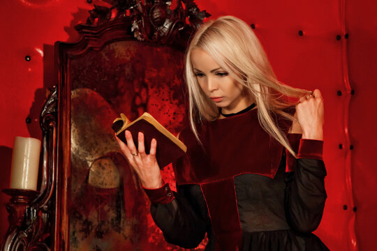 Queen of fear night Halloween, pensive blonde lady in black reading old book sitting in blood red room with abandoned mirror in medieval castle. Image of Witchcraft horror concept. Copy ad text space