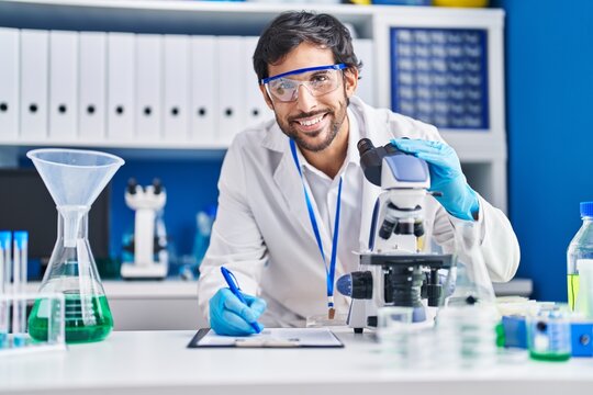 Young hispanic man scientist using microscope write on clipboard at laboratory
