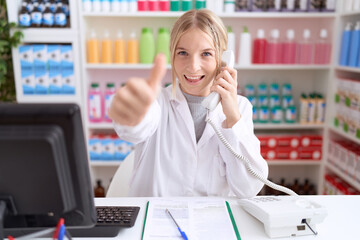 Young caucasian woman working at pharmacy drugstore speaking on the telephone approving doing positive gesture with hand, thumbs up smiling and happy for success. winner gesture.