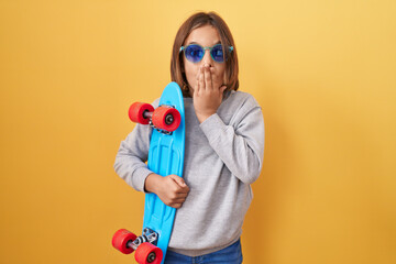 Little hispanic boy wearing sunglasses holding skate covering mouth with hand, shocked and afraid...