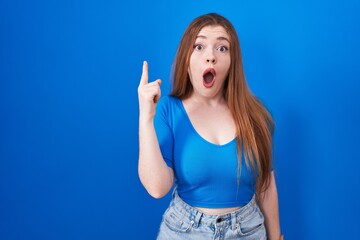 Redhead woman standing over blue background pointing finger up with successful idea. exited and happy. number one.