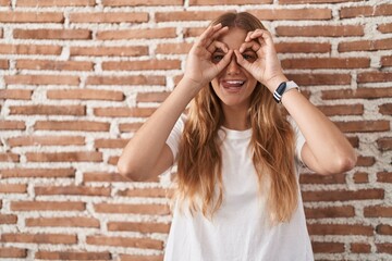 Young caucasian woman standing over bricks wall doing ok gesture like binoculars sticking tongue...