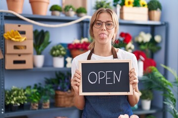 Young caucasian woman working at florist with open sign sticking tongue out happy with funny expression.