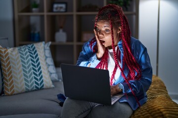 African american woman with braided hair using computer laptop at night afraid and shocked, surprise and amazed expression with hands on face