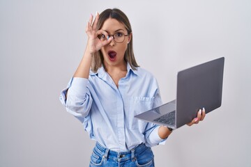 Young woman working using computer laptop doing ok gesture shocked with surprised face, eye looking through fingers. unbelieving expression.