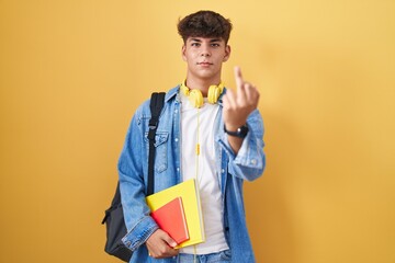Hispanic teenager wearing student backpack and holding books showing middle finger, impolite and...