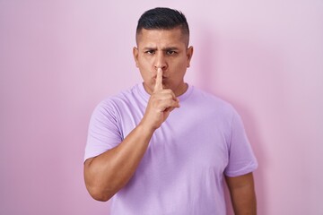 Young hispanic man standing over pink background asking to be quiet with finger on lips. silence and secret concept.