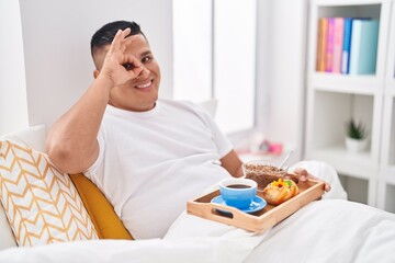 Young hispanic man eating breakfast in the bed smiling happy doing ok sign with hand on eye looking through fingers