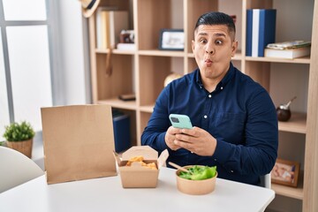 Hispanic young man eating take away food using smartphone making fish face with mouth and squinting eyes, crazy and comical.