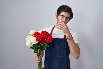 Young hispanic man holding bouquet of white and red roses bored yawning tired covering mouth with hand. restless and sleepiness.