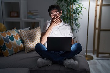 Young hispanic man using laptop at home at night laughing and embarrassed giggle covering mouth with hands, gossip and scandal concept