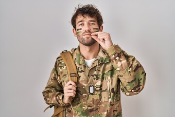 Hispanic young man wearing camouflage army uniform mouth and lips shut as zip with fingers. secret...