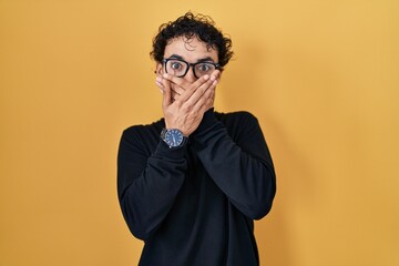 Hispanic man standing over yellow background shocked covering mouth with hands for mistake. secret...