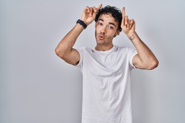 Hispanic man standing over isolated background doing funny gesture with finger over head as bull horns