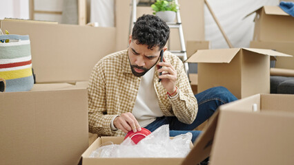 Young hispanic man talking on smartphone unpacking cardboard box at new home