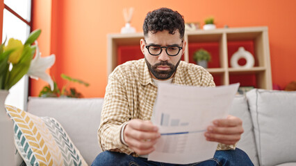 Young hispanic man reading document sitting on sofa at h