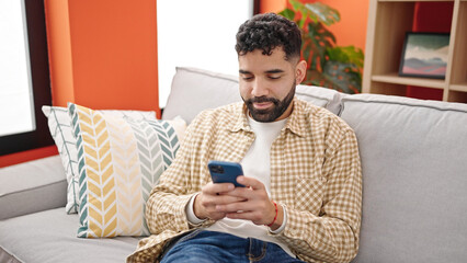Young hispanic man using smartphone sitting on sofa at h