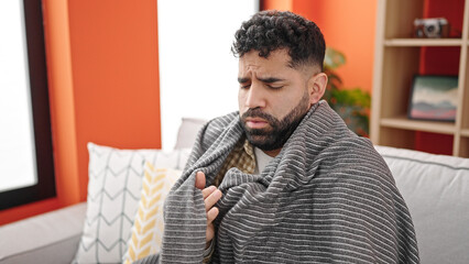 Young hispanic man sitting on sofa covering with blanket for cold at h