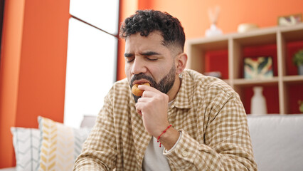 Young hispanic man eating doughnut sitting on sofa at h