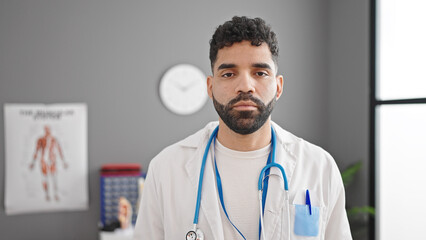 Young hispanic man doctor standing with serious expression at clinic