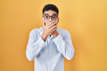 Hispanic man with beard standing over yellow background shocked covering mouth with hands for...