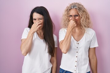 Mother and daughter standing together over pink background smelling something stinky and disgusting, intolerable smell, holding breath with fingers on nose. bad smell - obrazy, fototapety, plakaty