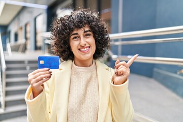 Young brunette woman with curly hair holding credit card smiling happy pointing with hand and...