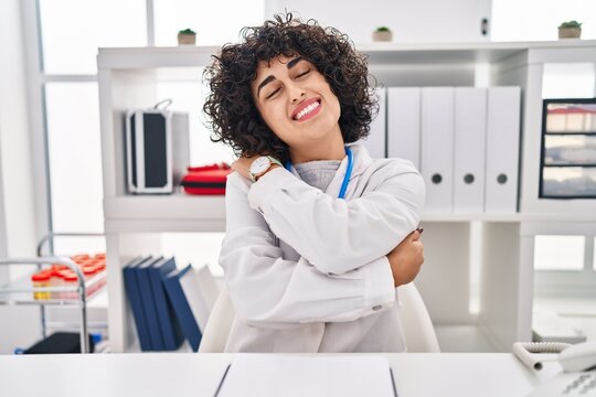 Young brunette woman with curly hair wearing doctor uniform and stethoscope hugging oneself happy and positive, smiling confident. self love and self care