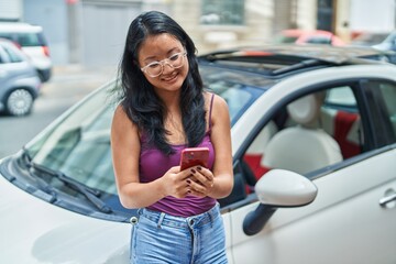 Young chinese woman using smartphone leaning on car at street