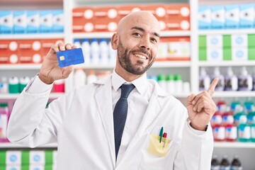 Middle age bald man working at pharmacy drugstore holding credit card smiling happy pointing with hand and finger to the side