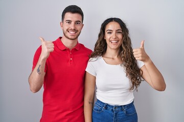 Young hispanic couple standing over isolated background doing happy thumbs up gesture with hand. approving expression looking at the camera showing success.
